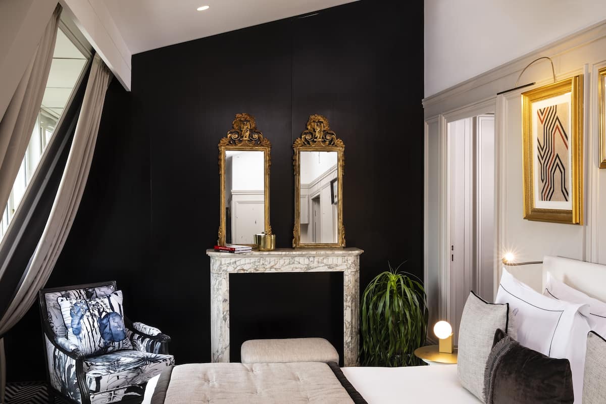 A modern bedroom with a black wall, a marble fireplace, two ornate mirrors, a patterned armchair, a bedside table with a lamp, and a white bed with pillows.