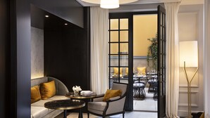 Una habitación de hotel moderna con sofá, sillón y una mesa redonda. Se ve un gran ventanal con cortinas y vistas al comedor.