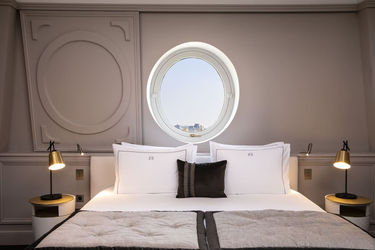 A hotel room with a large bed, two bedside tables, a round mirror above the bed, and a view of the city through the window.