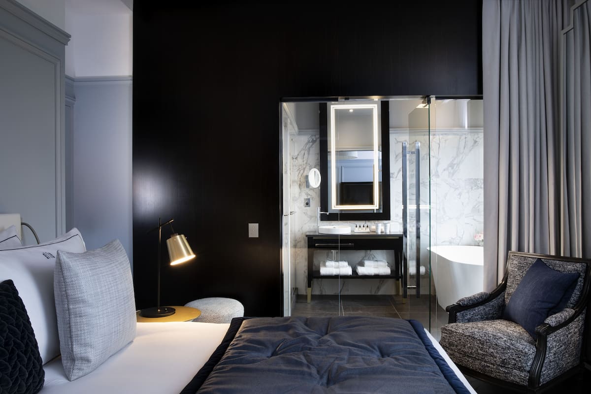 A modern hotel room with a large bed, a sitting area with a chair, a small round table, a bedside lamp, a bathroom with a mirror, and a vanity with a sink.