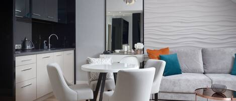 Eine moderne Küche mit weißen Schränken, einem runden Esstisch und weißen Stühlen. Im Wohnbereich befindet sich eine graue Couch mit blau und orangefarbenen Kissen.