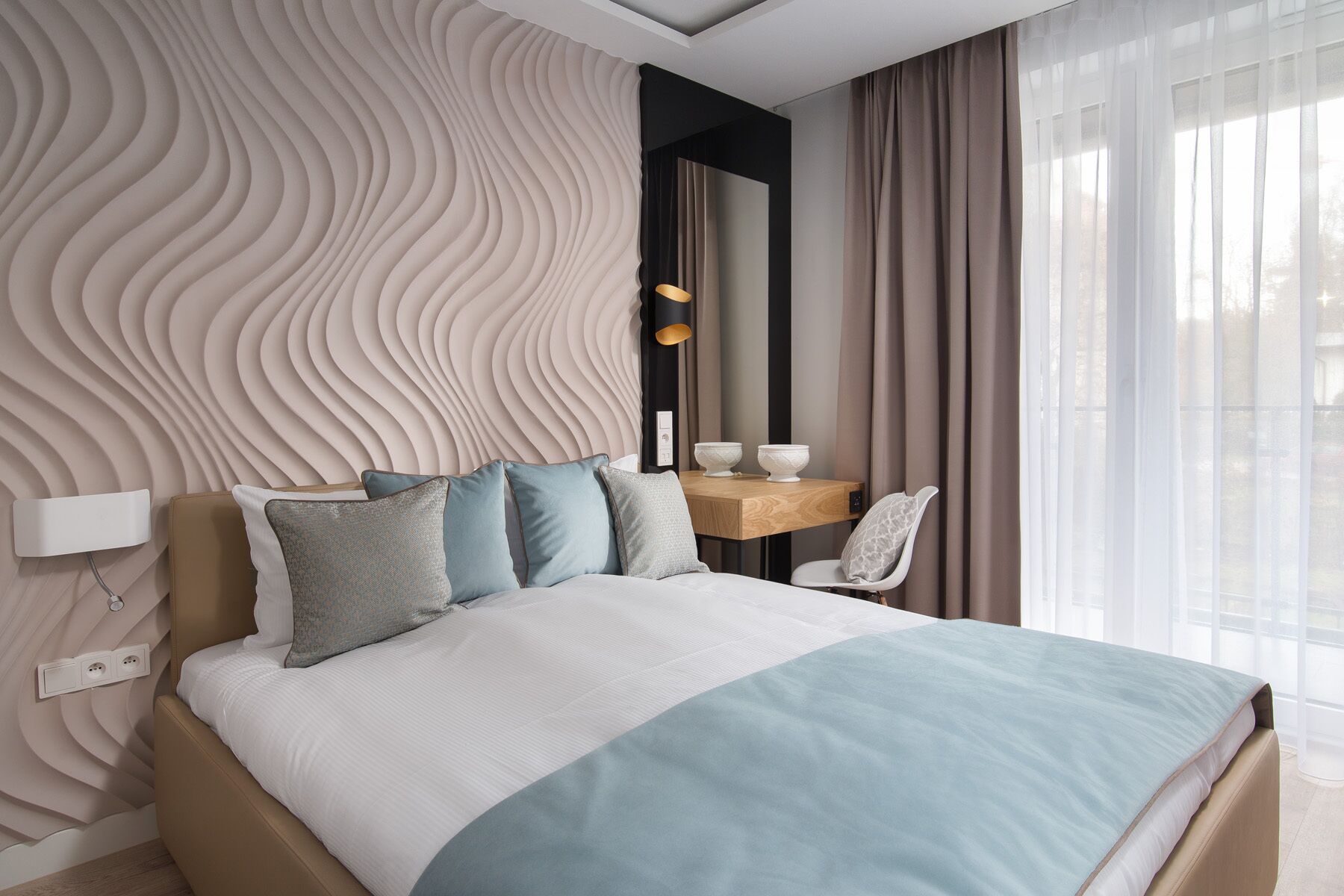 Ein modernes Schlafzimmer mit einem Kopfende im Wellenmuster, einem Holfnachtkästchen mit zwei Schalen, einem weißen Stuhl und einem großen Fenster mit transparenten Vorhängen.