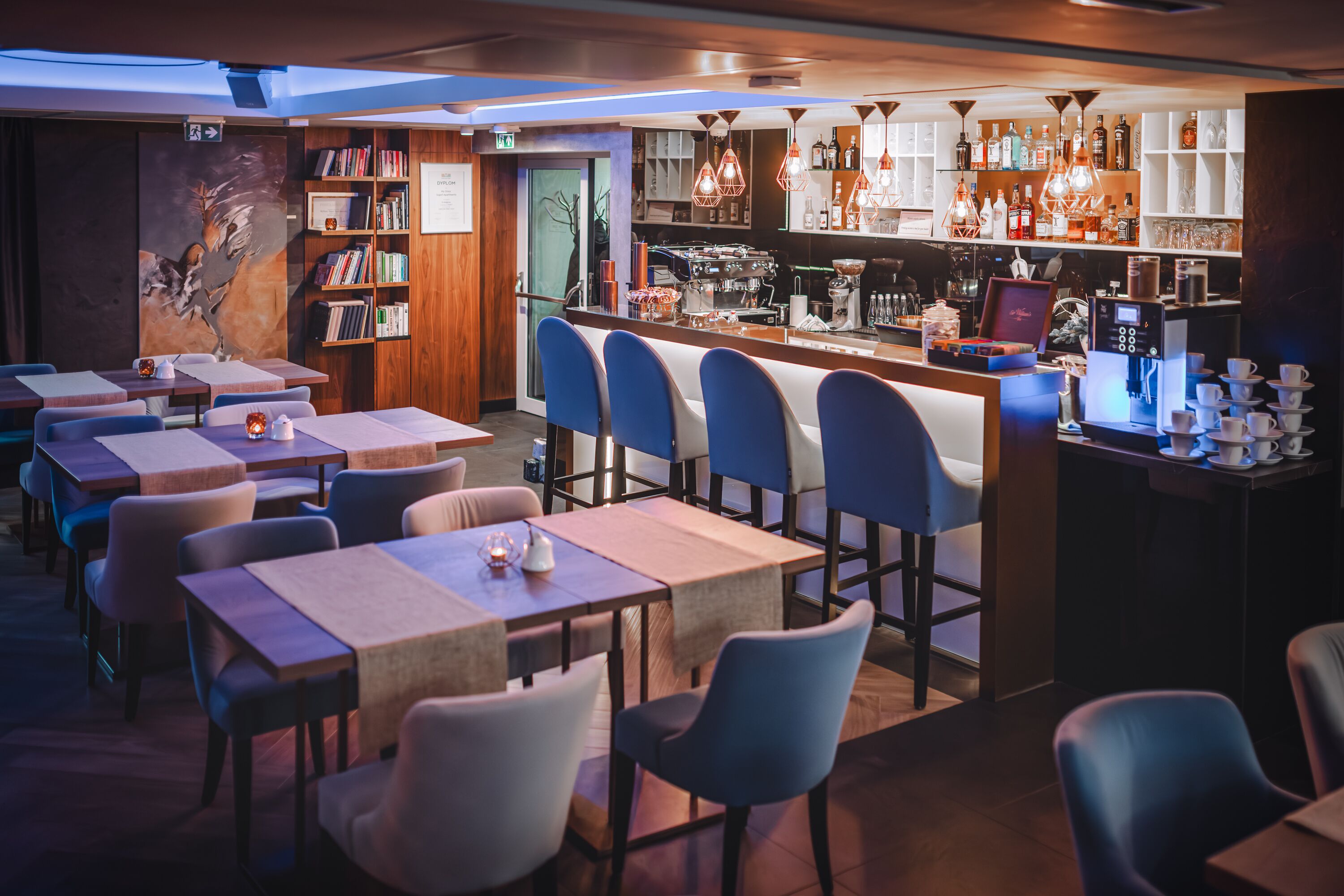 A restaurant with multiple tables and chairs, a bar area with a coffee machine, and a bookshelf with books.