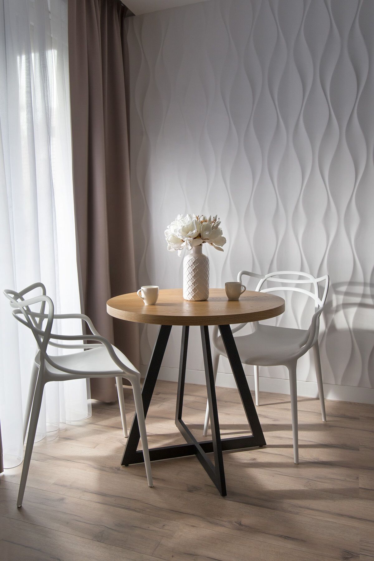 A round wooden table with a white vase and two cups, flanked by two white chairs with black legs, set against a patterned wall and a sheer curtain.