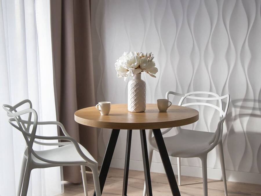 A round wooden table with a white vase and two cups, flanked by two white chairs with black legs, set against a patterned wall and a sheer curtain.