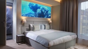 A modern hotel room with a large bed, a wall-mounted TV displaying an underwater scene, bedside lamps, and a view of the outside through a window with curtains.