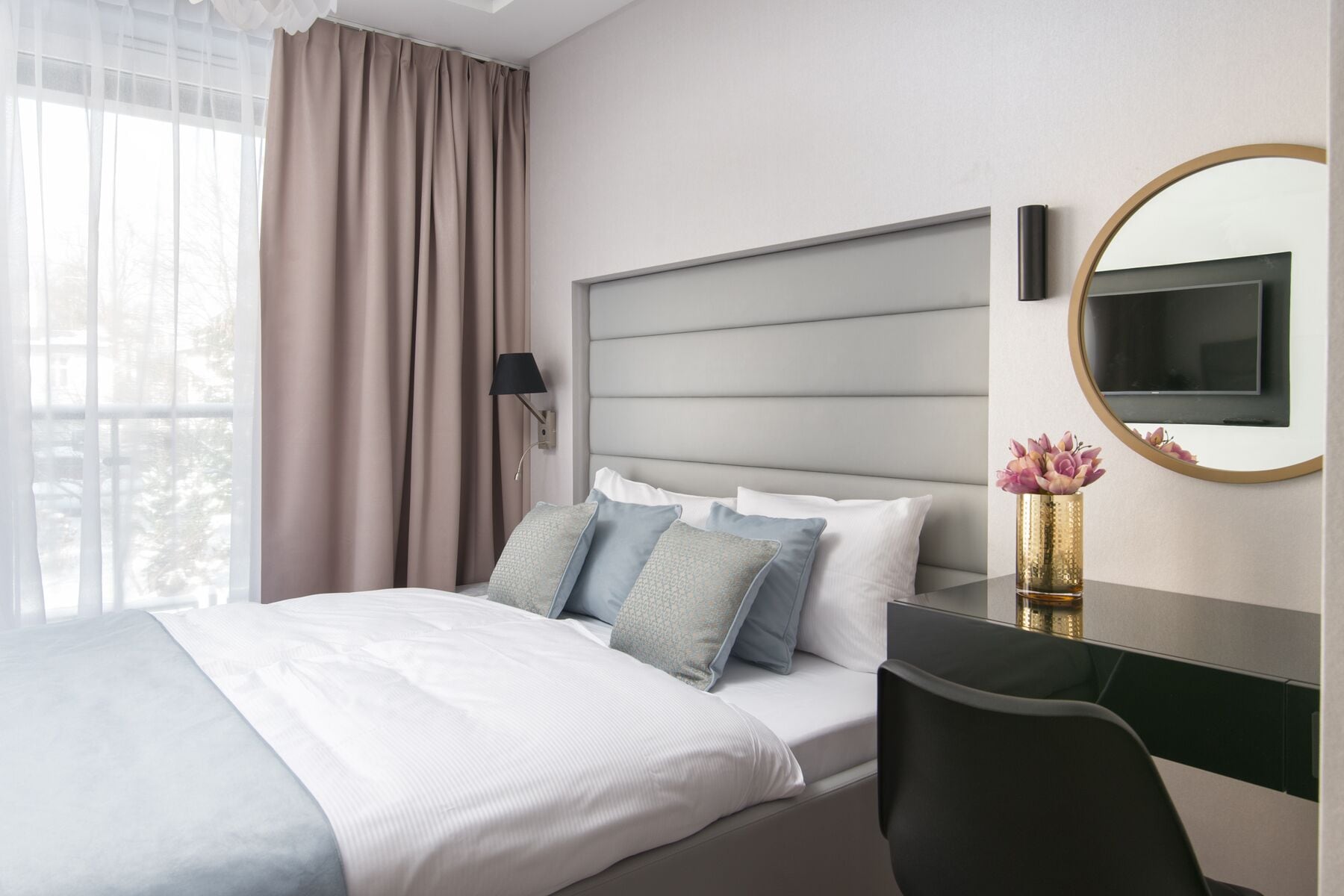A modern hotel room with a large bed, a desk, a chair, a TV, and a mirror.