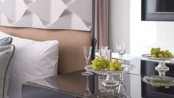 A modern hotel room with a dining table set for two, featuring a glass top and a decorative centerpiece of green grapes.