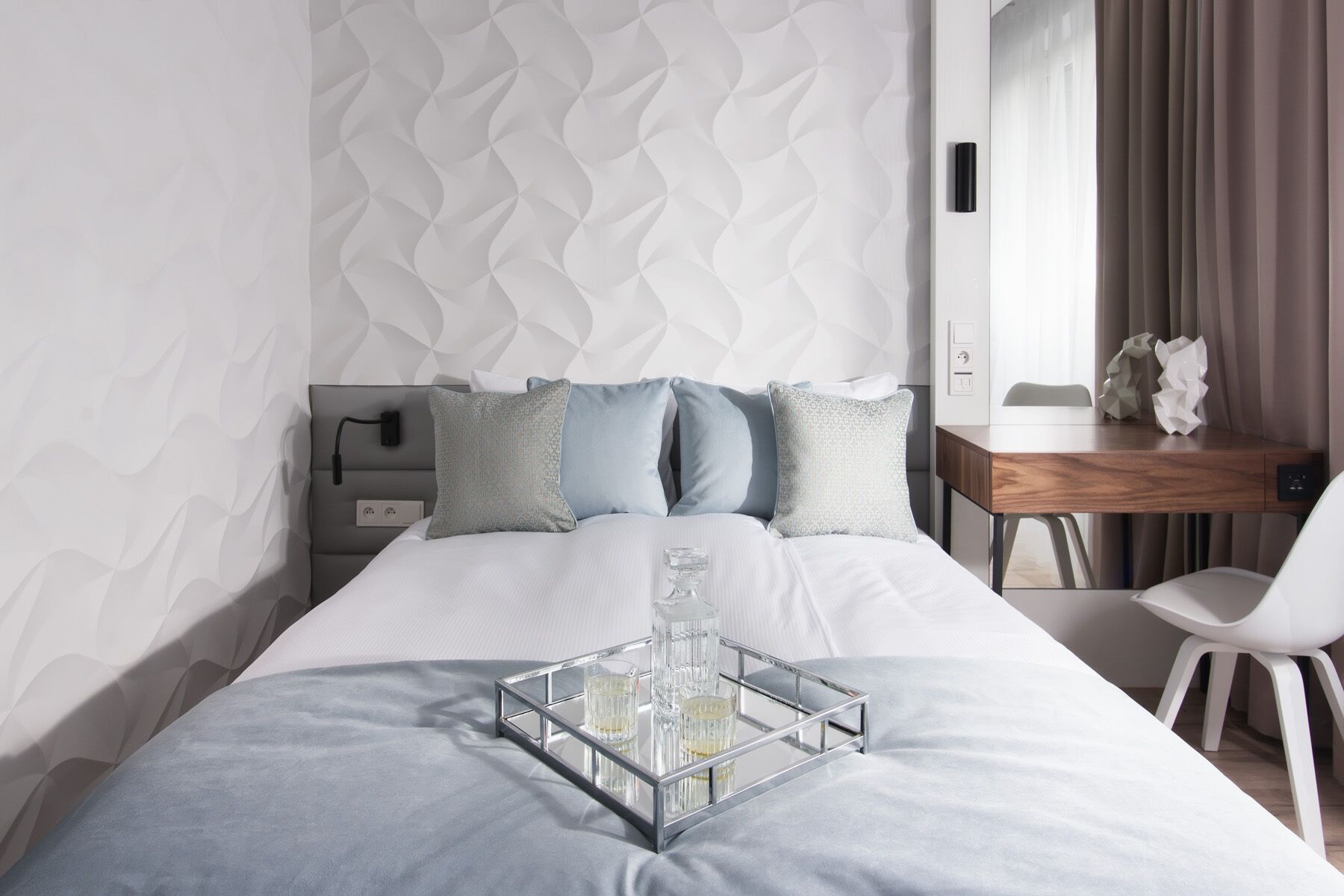 A modern hotel room with a bed, bedside table, and a desk with a chair.