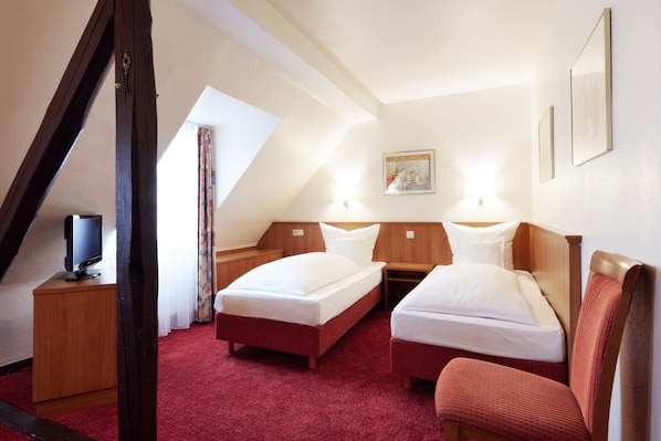 Habitación de hotel con dos camas individuales, una mesita de noche de madera, un televisor y una alfombra roja.