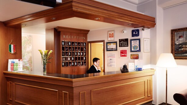 A reception desk with a wooden finish, a man standing behind it, and a wall with framed certificates and a map. - Hotel Doria (Düsseldorf)