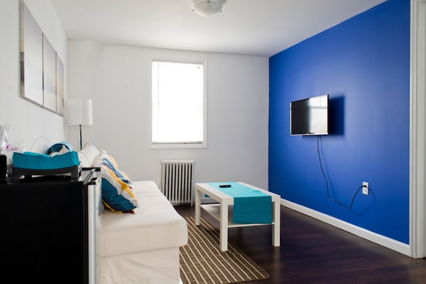 A modern living room with a white sofa, a small table, a blue accent wall, a mounted TV, and a window providing natural light.