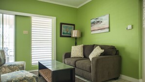 A living room with a green wall, a brown sofa, a glass coffee table, and a patterned armchair. - Breezy Shores at Cardiff Hall (Runaway Bay)