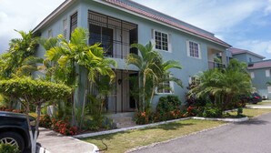 A two-story residential building with a tiled roof, large windows, and a well-maintained garden with palm trees and flowering plants. - Wellington Cove (Kingston)