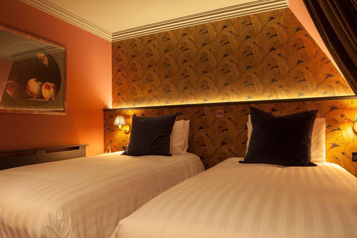 A hotel room with two beds, a wall-mounted lamp, and a painting of fruit.