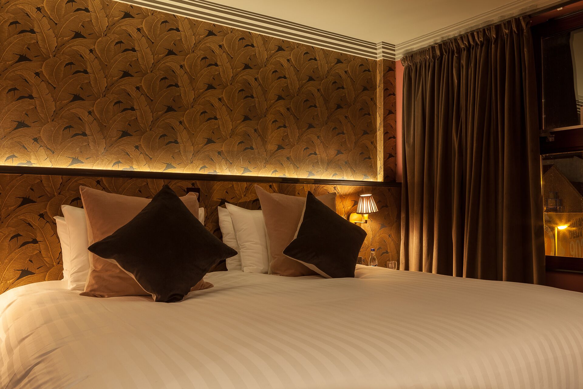 A hotel room with a large bed, multiple pillows, a wall with a feather pattern, a lamp, and a window with curtains.