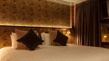 A hotel room with a large bed, multiple pillows, a wall with a feather pattern, a lamp, and a window with curtains.