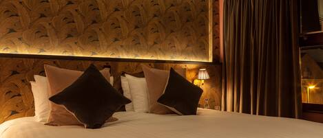 Hotelkamer met een groot bed, meerdere kussens, een muur met een verenmotief, een lamp en een raam met gordijnen.