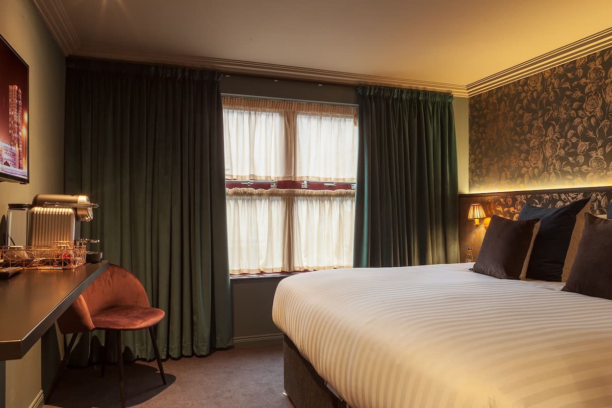 A hotel room with a bed, a desk with a chair, a window with curtains, and a wall with a floral pattern.