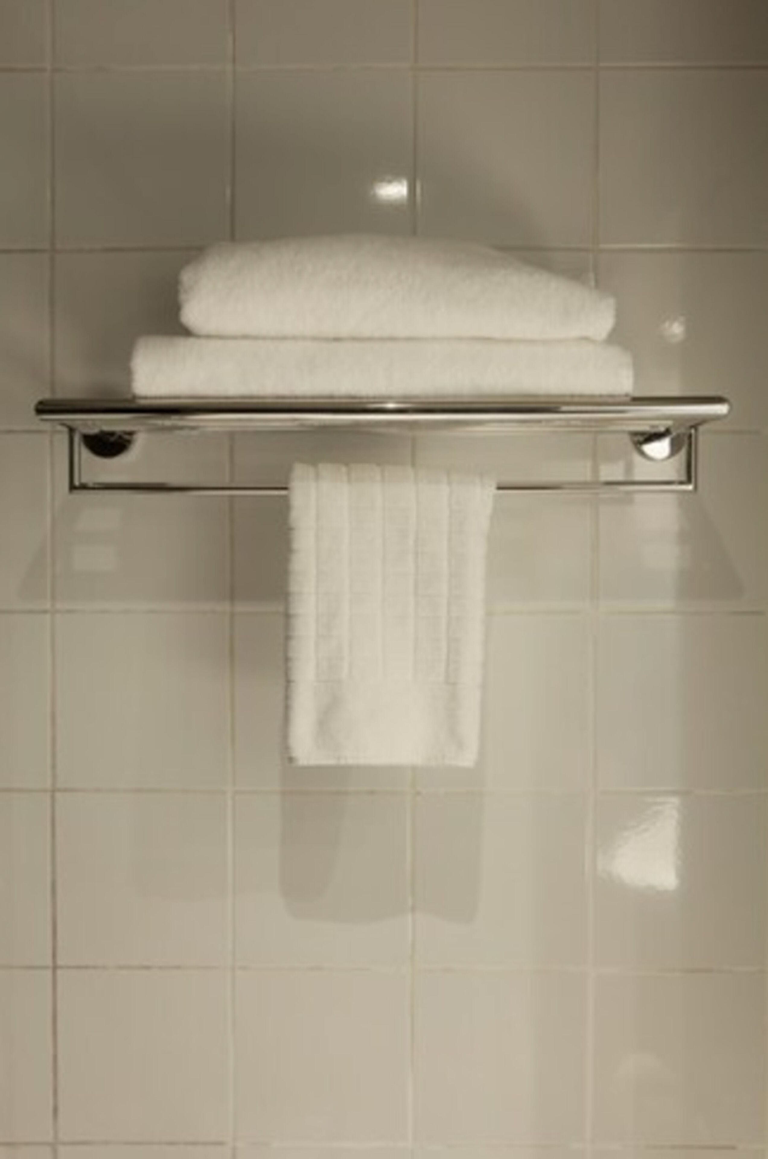 A bathroom with a white tiled wall, a metal towel rack holding two folded towels, and a single towel hanging from the rack.