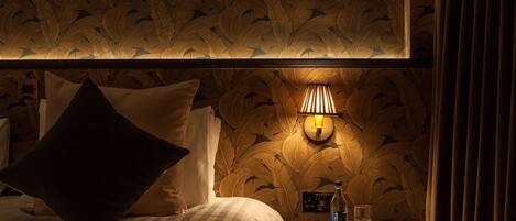 A hotel room with a bed, pillows, a wall-mounted lamp, a nightstand with a bottle and glass, and a patterned wallpaper.