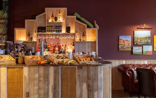 Una zona de bar con estantes de madera que muestran botellas y vasos, una máquina de café, y diversos aperitivos y frutas.