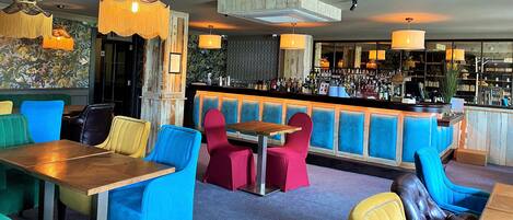 Zona de bar con sillas de colores y una mesa de madera, barra con diferentes botellas y una lámpara decorativa en el techo.