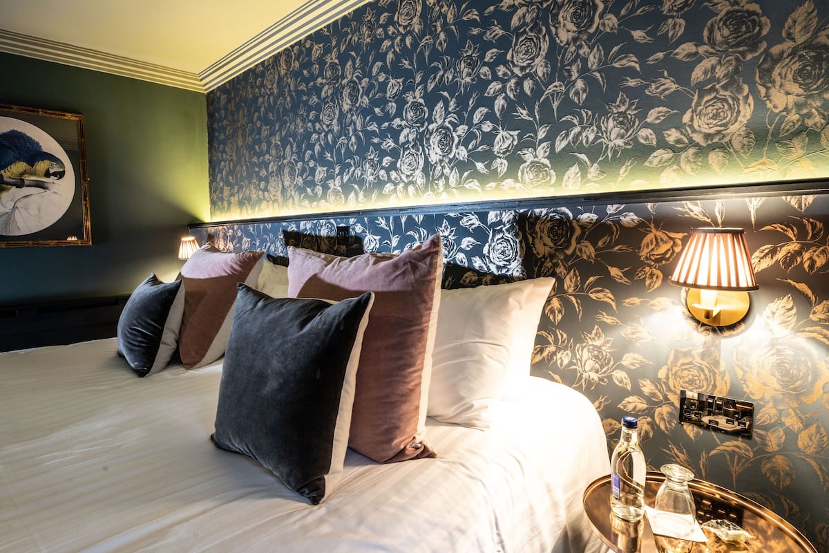 A hotel room with a large bed, multiple pillows, a nightstand with a lamp, and a floral wallpaper.