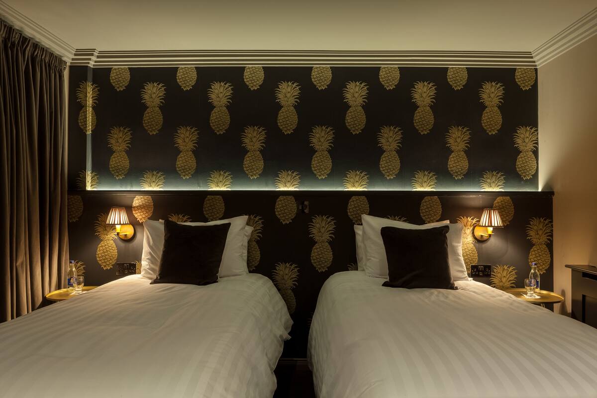 A hotel room with two beds, a dark pineapple-patterned headboard, and wall-mounted lamps.