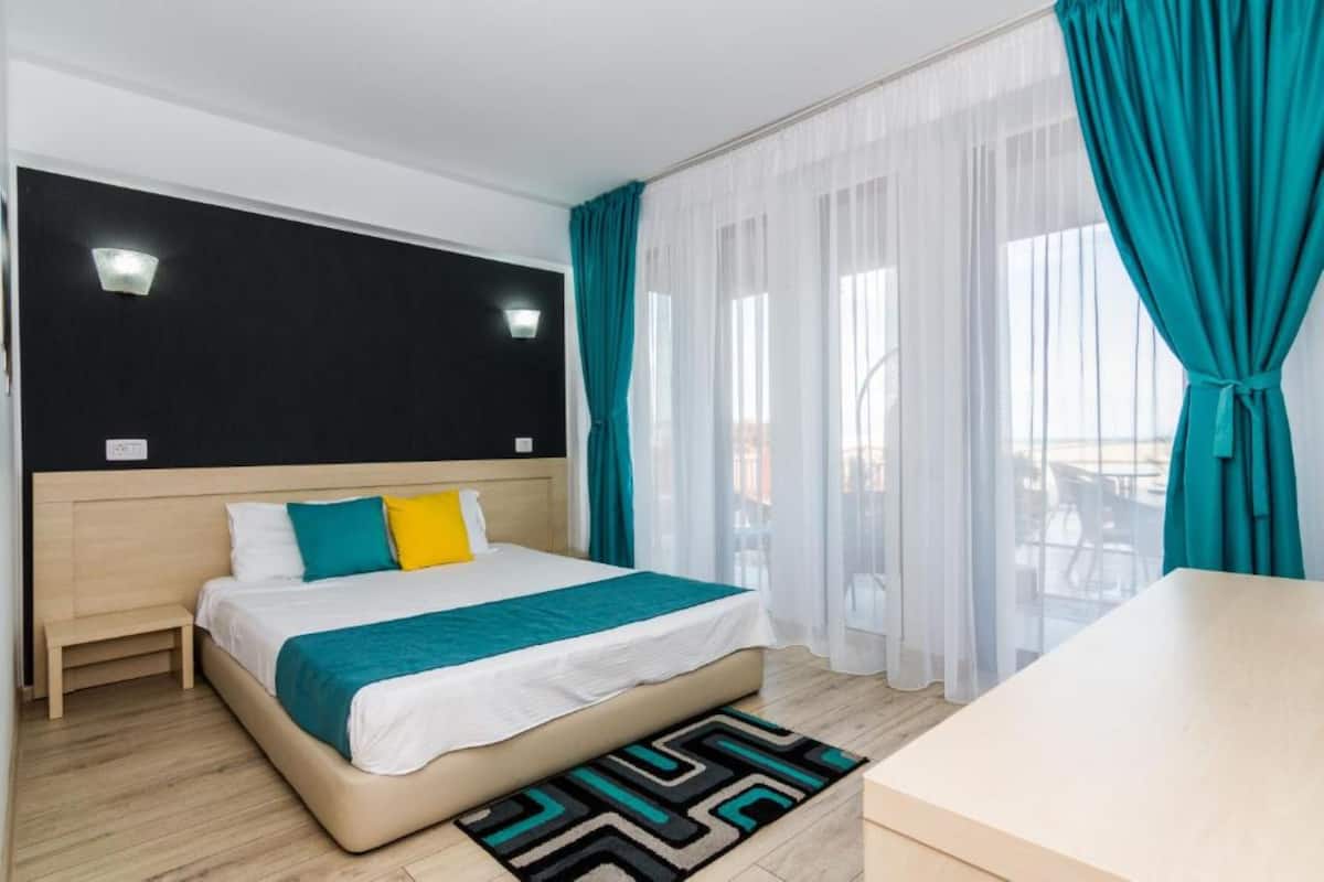 Ein modernes Hotelzimmer mit Bett, Nachttisch, Schreibtisch und Blick ins Freie durch transparente Vorhänge.