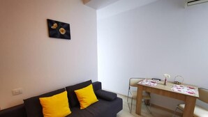 Un salon avec un canapé foncé, deux coussins jaunes, une table à manger en bois recouverte d’une nappe à motifs floraux et une œuvre d’art accrochée au mur.
