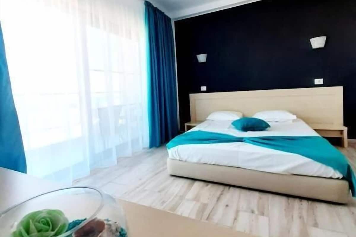 Ein Hotelzimmer mit einem Bett, einem Nachttisch, einem Fenster mit Vorhängen und Blick ins Freie.