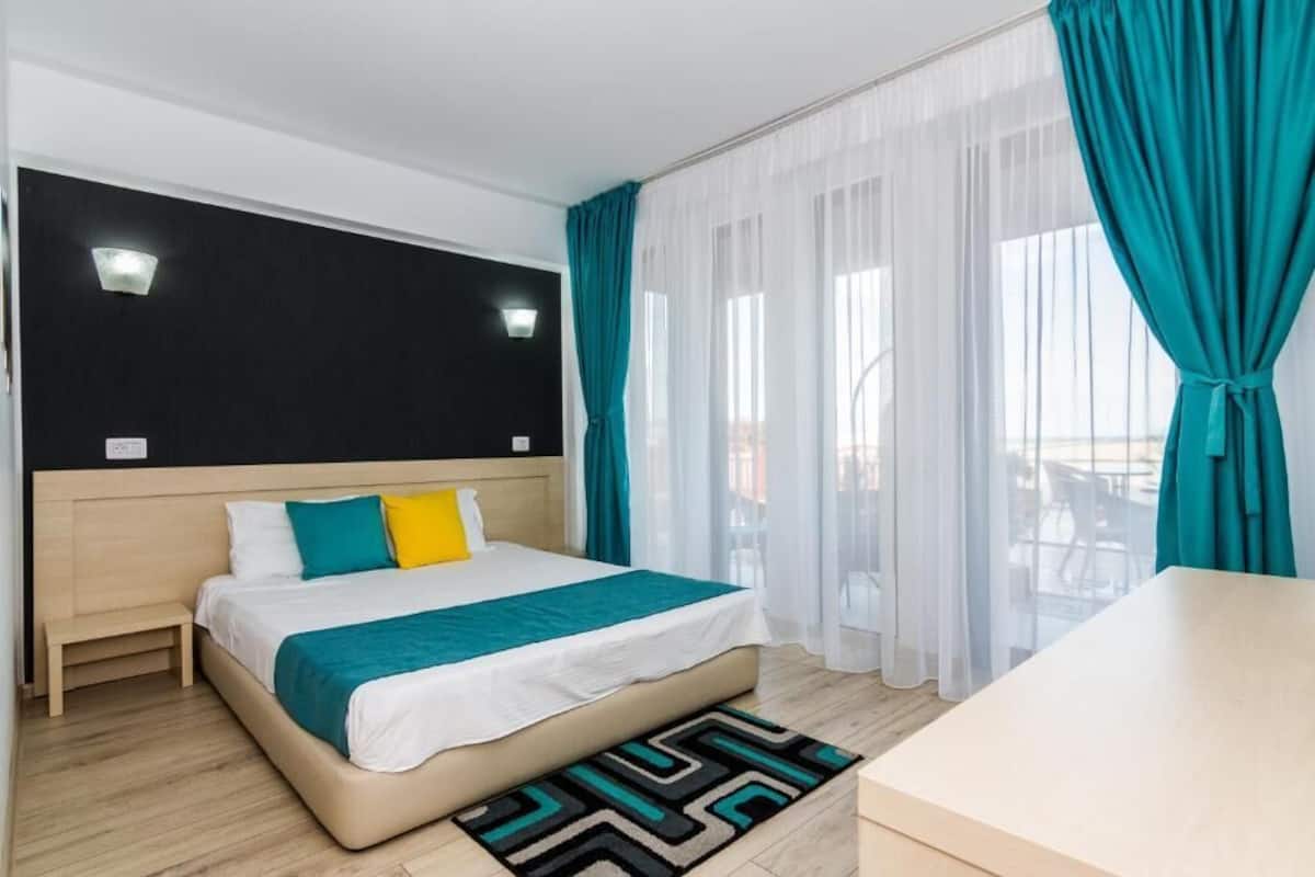 Ein Hotelzimmer mit Bett, Nachttisch, Schreibtisch und Blick durch transparente Vorhänge nach draußen.
