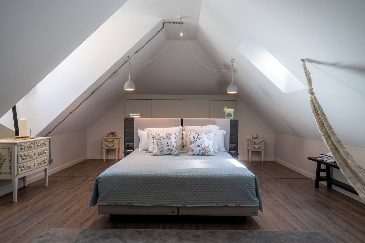 Een slaapkamer met een schuine kap, een bed met bloemenkussens, twee nachtkastjes en een hangmat.