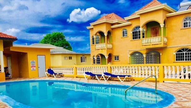 A swimming pool with lounge chairs, a tiled deck, and a yellow building with arched windows and balconies. - Eagles Nest (Montego Bay)