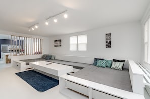 A modern living room with a large sectional sofa, a white coffee table, and a blue rug. There are two windows with blinds, a kitchen area in the background, and a ceiling track lighting system.