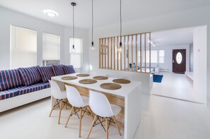 A modern living room with a white sofa, a dining table with four chairs, and a kitchen area in the background.