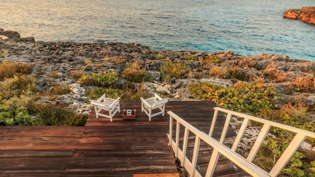 A wooden deck with two white chairs and a small table, overlooking a rocky shoreline and calm sea. - Paradise  Beach Front Apartment (Discovery Bay)