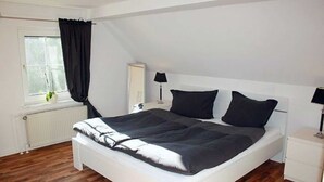 Une chambre à coucher avec un grand lit, deux tables de chevet blanches, une fenêtre avec des rideaux et un radiateur.