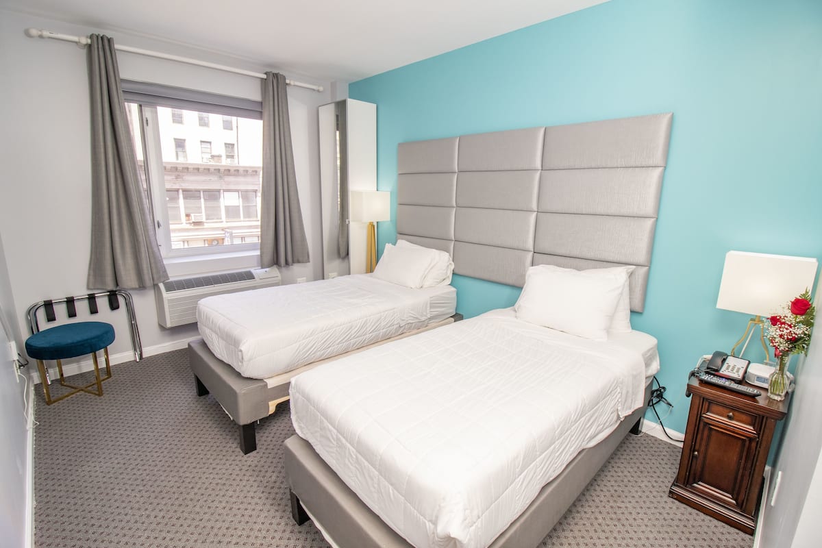 A hotel room with two beds, a window with curtains, a small table with a lamp, and a wall-mounted air conditioning unit.