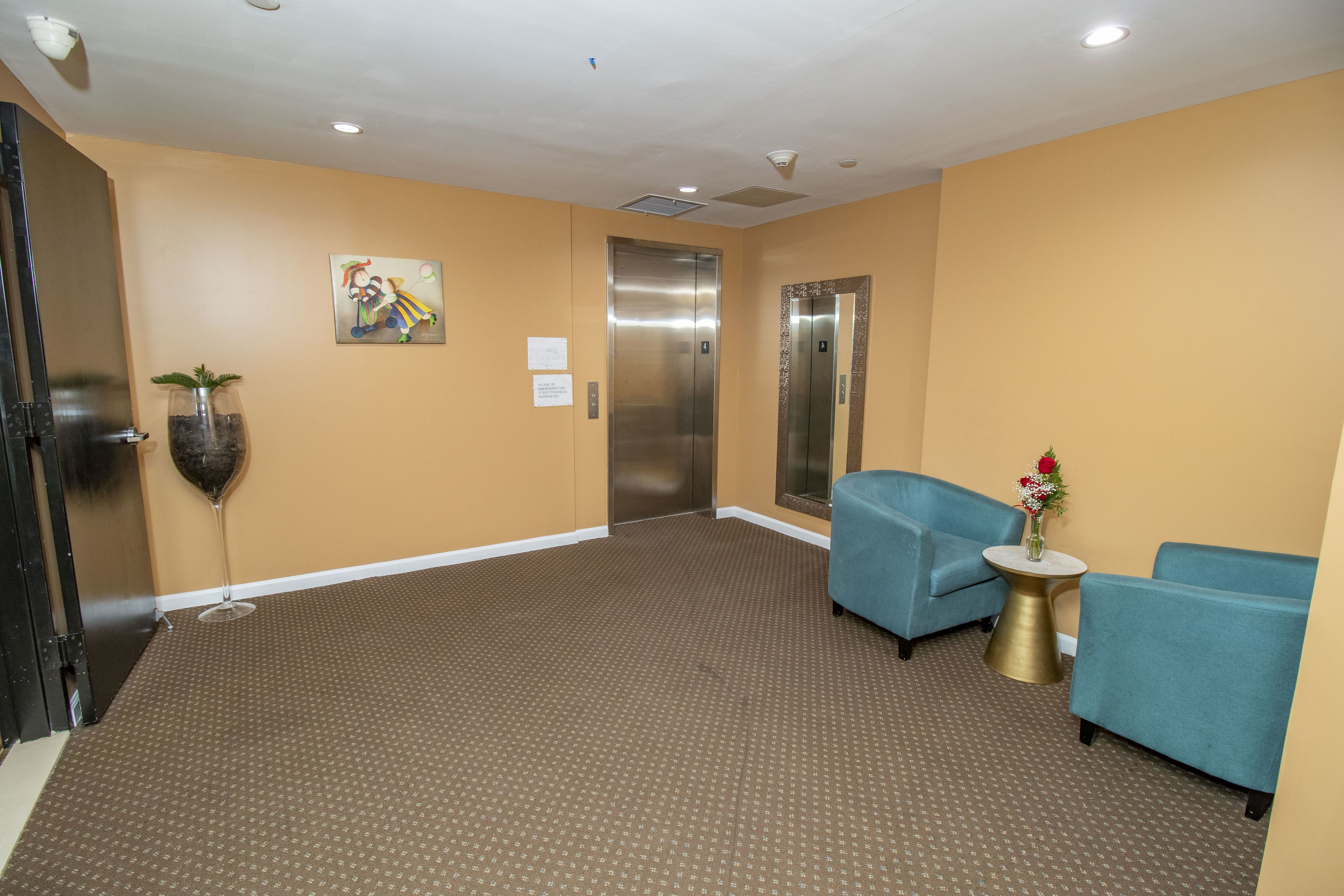 A hotel lobby with a brown patterned carpet, two blue armchairs, a small round table with a vase of flowers, and an elevator.