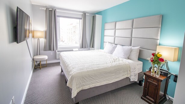 A hotel room with a large bed, a bedside table with a lamp, a TV mounted on the wall, and a window with curtains. - Canal Loft Hotel (New York)