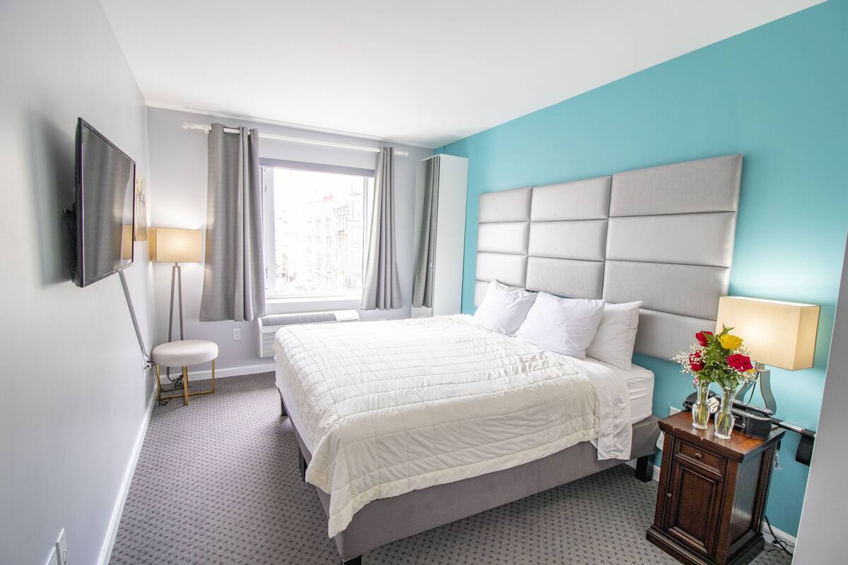 A hotel room with a large bed, a bedside table with a lamp, a TV mounted on the wall, and a window with curtains.