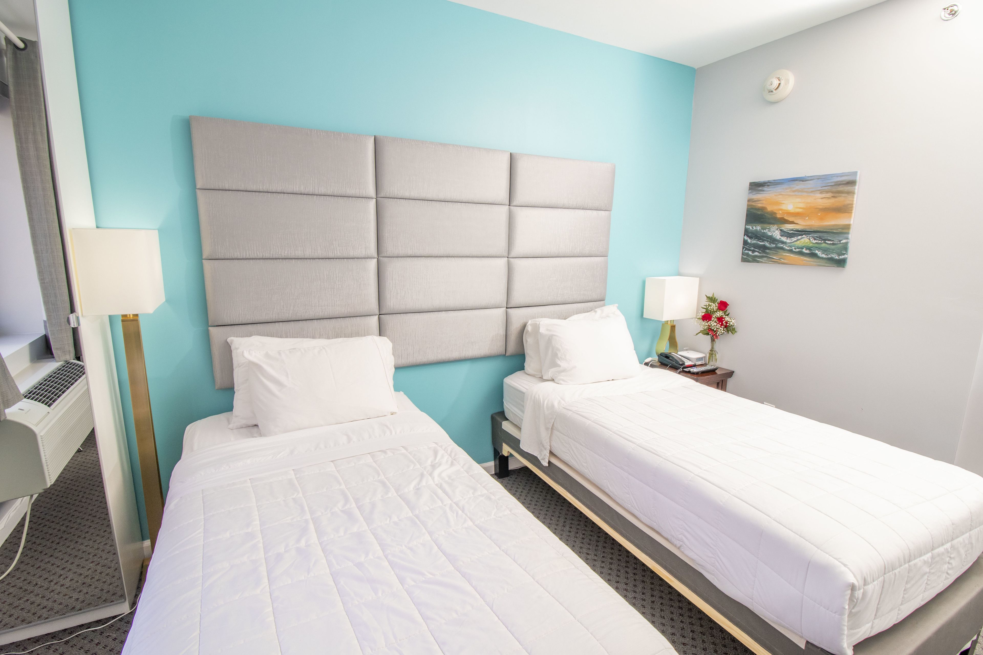 Habitació d'hotel amb dos llits individuals, un capçal gran i amb folrar, un escriptori amb una làmpada i una taula petita amb un ram de flors.
