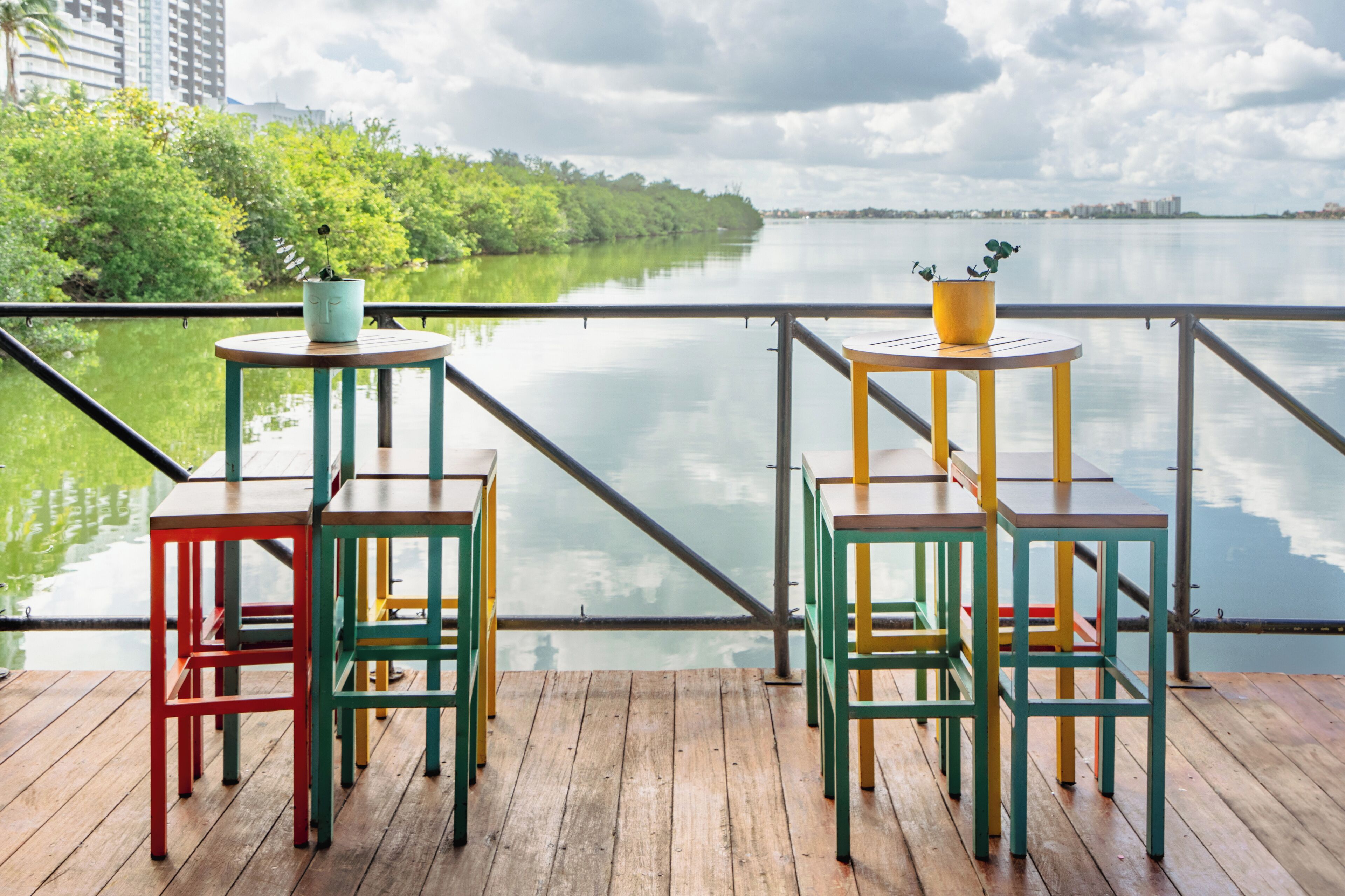 A deck with colorful chairs and tables overlooking a calm body of water, with greenery and buildings in the background.