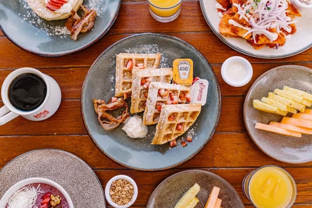Una mesa de desayuno con waffles, tocino y diferentes salsas.
