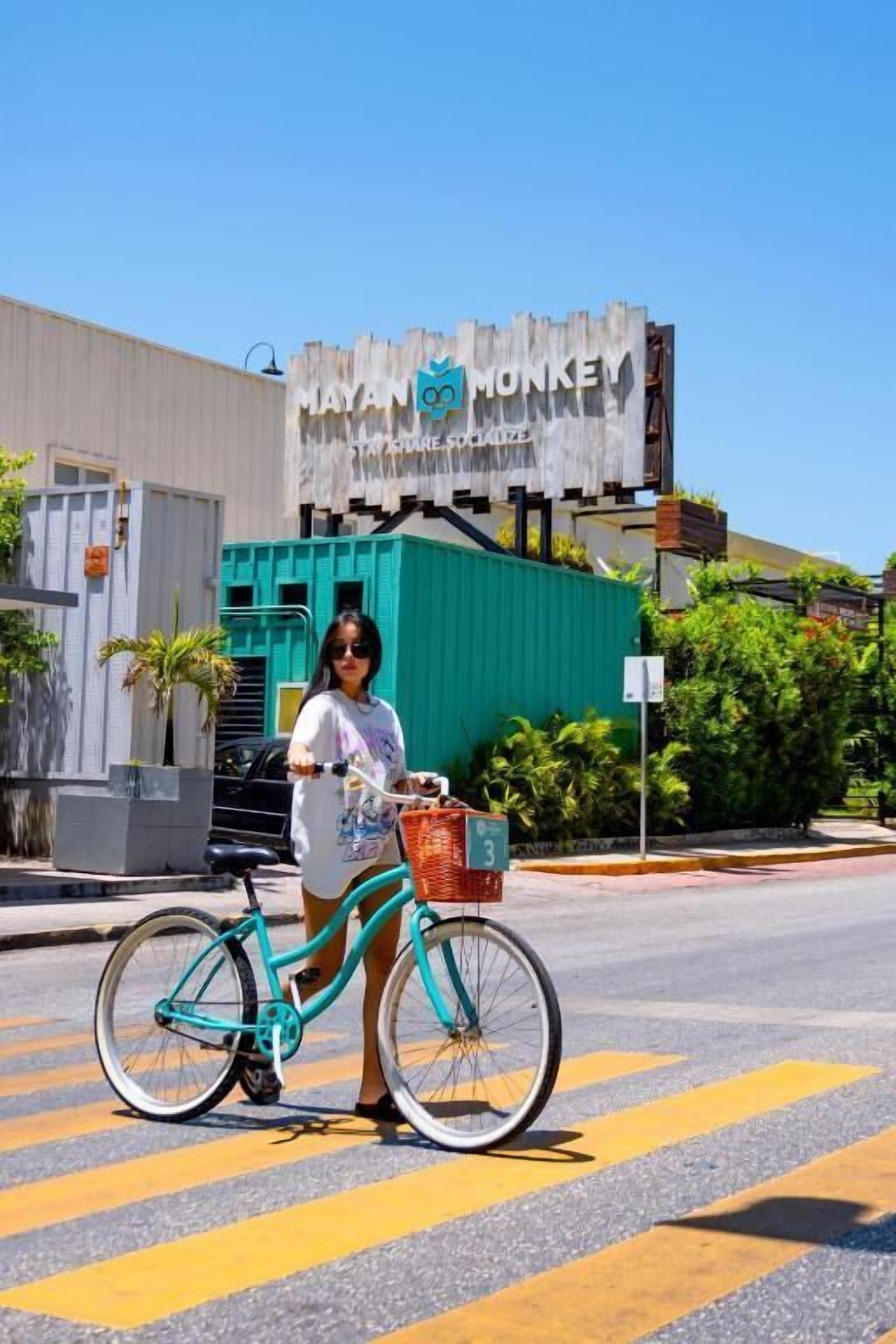 Una persona de pie junto a una bicicleta turquesa con una canasta, frente a un edificio con un letrero que dice "MAYAN MONKEY".