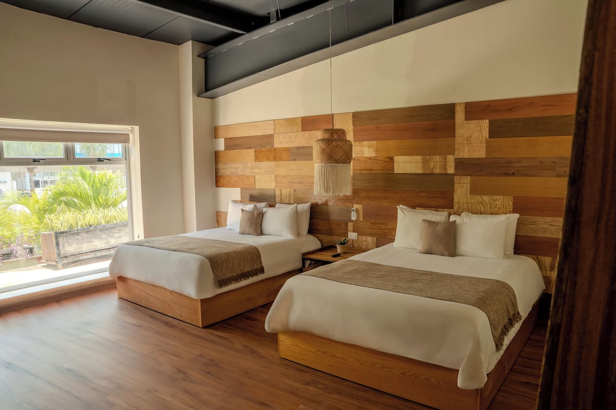 A hotel room with two beds, wooden headboards, a large window with a view, and a hanging light fixture.