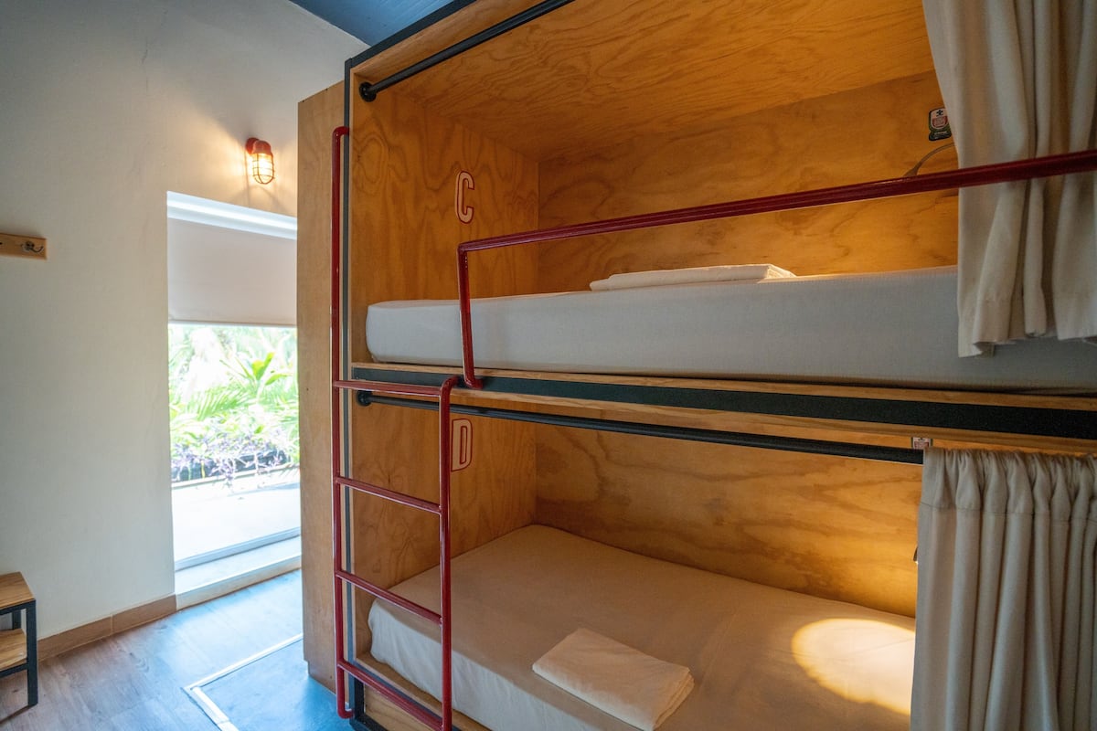 A bunk bed with a wooden headboard, a small table, and a window with a view of greenery.