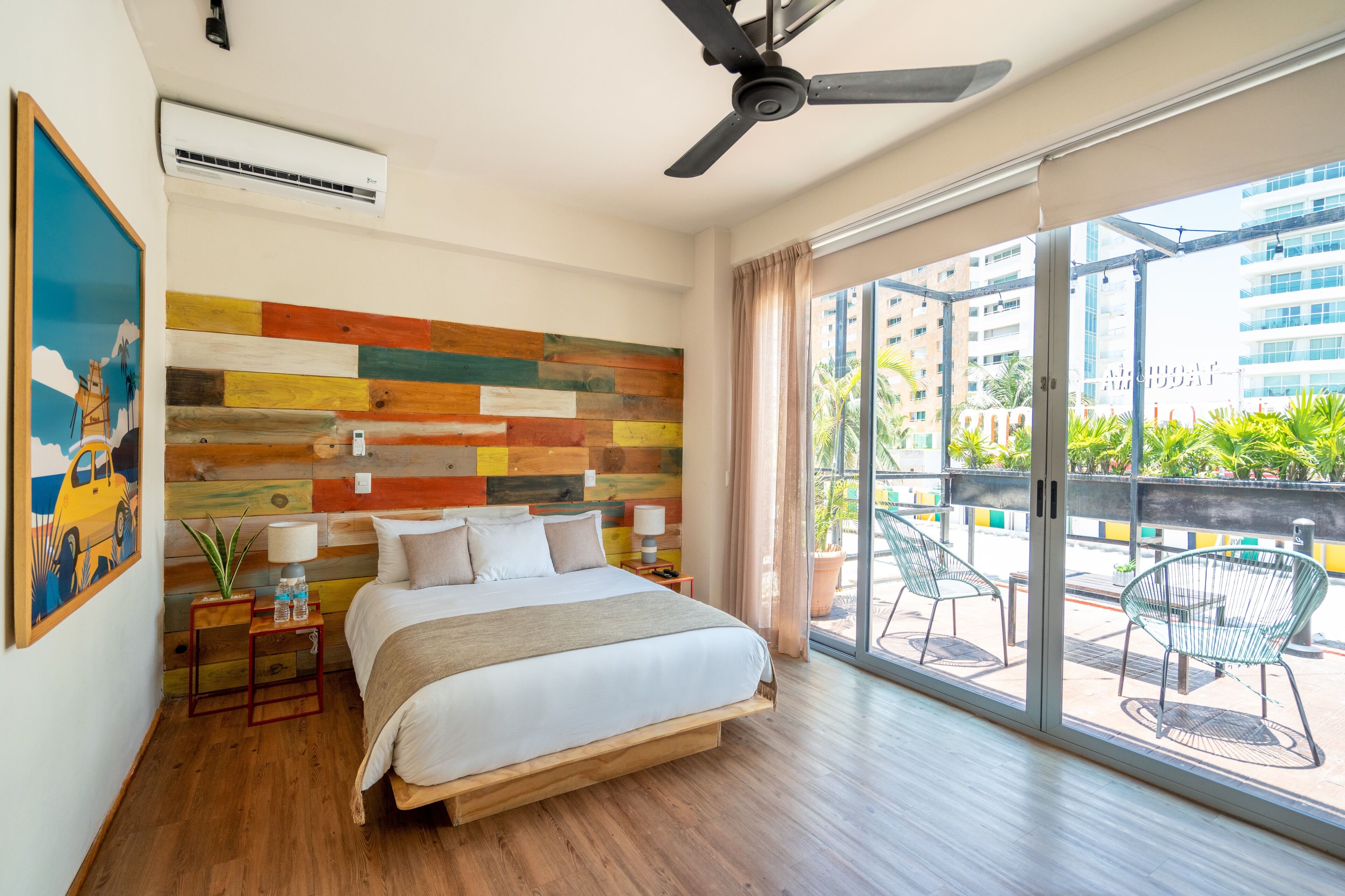 A modern hotel room with a large bed, a ceiling fan, a colorful wooden wall, a large window with a view of the city, and a balcony with chairs.